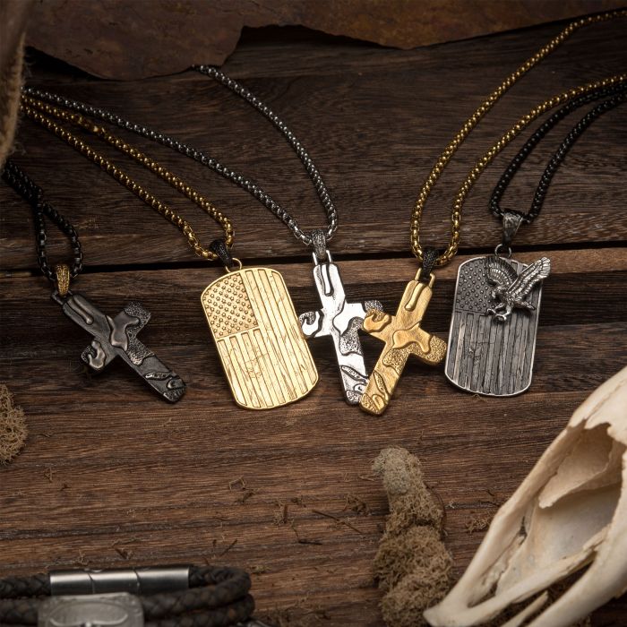 A variety of military-themed dog tag necklaces from INOX are displayed on a wooden surface. Each Stainless Steel Gun Metal Plated 3D Canyon Pattern Pendant features an embossed design, including a cross, the American flag, and an eagle. The tags hang from silver and gold chains, surrounded by rustic elements.