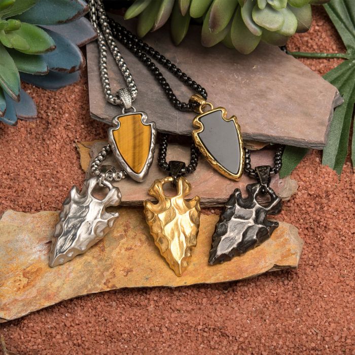 A collection of five arrowhead-shaped necklaces on a textured, reddish-brown surface with layered stones and green succulents around. The men's jewelry pieces vary in material and color, including the Stainless Steel Gun Metal Plated Chiseled Arrowhead Pendant | 24" from INOX, silver, gold, and patterned designs.