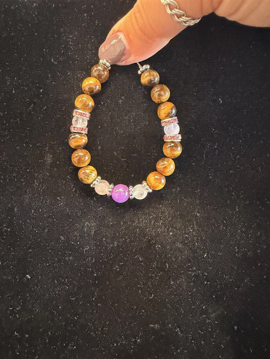 A hand holds the Redemption Jewelry B010 Tiger Eye, Rose Quartz & Amethyst Bracelet, featuring a mix of brown Tiger Eye, soft pink Rose Quartz, vivid amethyst beads, gold beads, silver spacers, and clear accents—displayed against a black background.