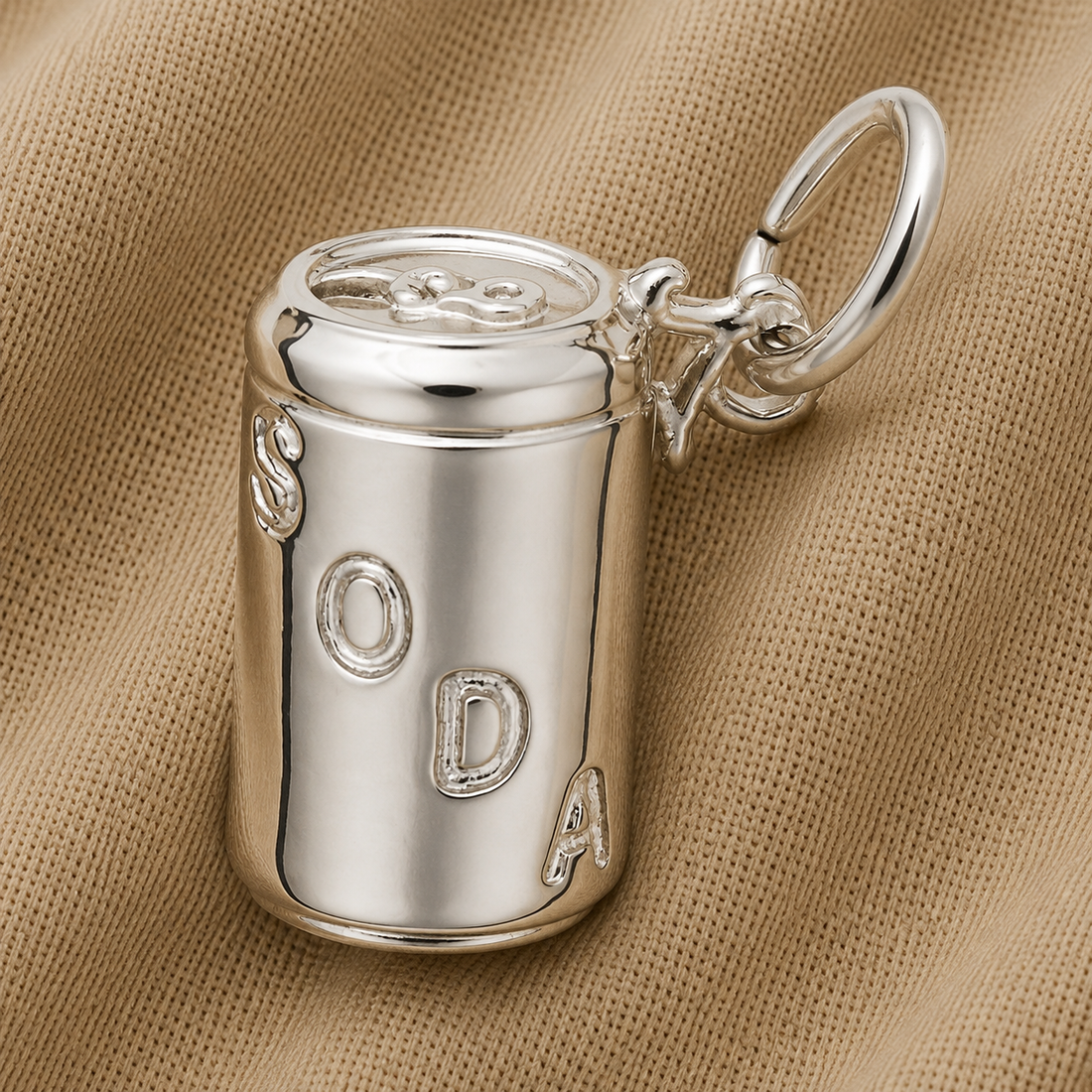 The Soda Can Charm—Sterling Silver by Rembrandt Charms is a three-dimensional, silver jewelry piece shaped like a soda can with “SODA” embossed on it, displayed on a textured beige fabric background.
