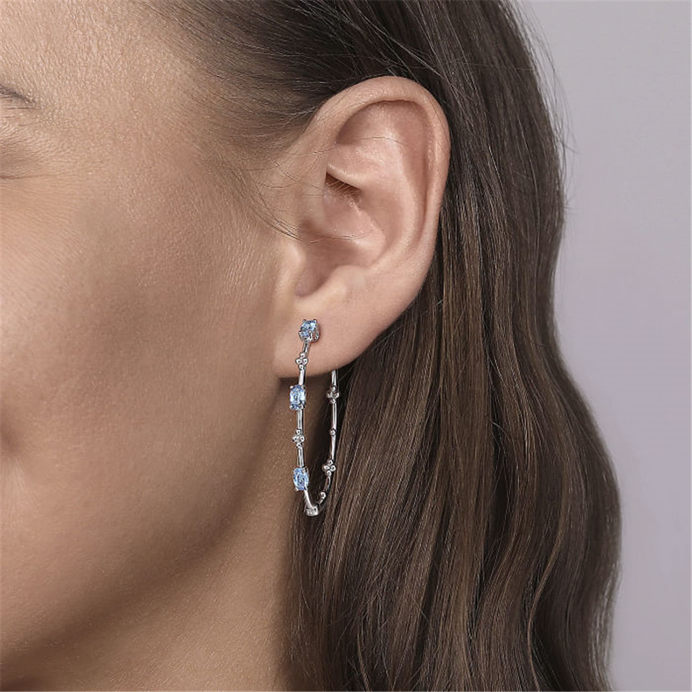 Close-up side view of a person wearing Gabriel & Co's 925 Sterling Silver Swiss Blue Topaz Bujukan Classic Hoop Earrings. The hoop earring, encrusted with radiant Swiss blue topaz gemstones, features a loop design that contours the earlobe, accentuating the sparkle of the gemstones against the skin. The background is neutral and blurred.