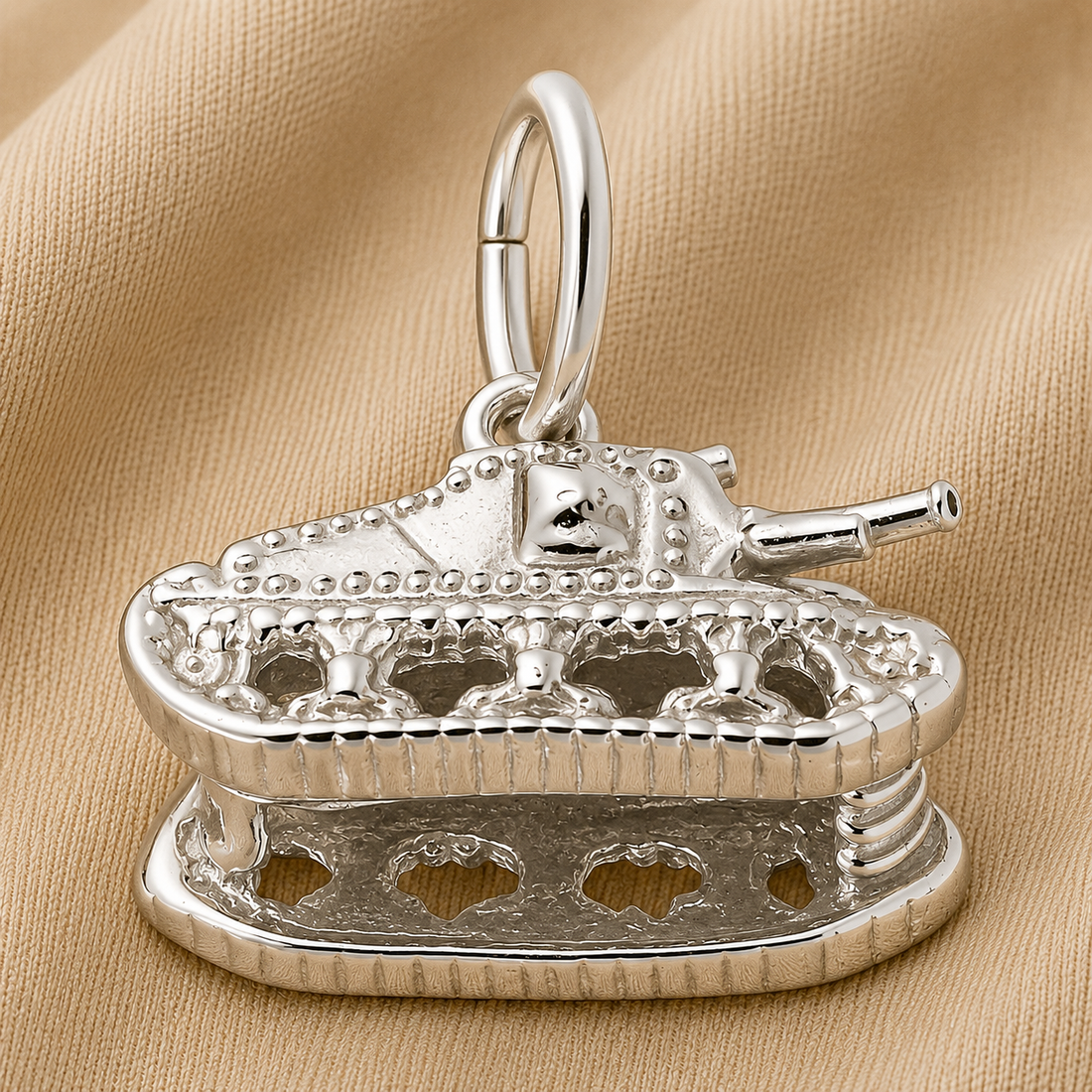 A Tank Charm- Sterling Silver by Rembrandt Charms rests on tan fabric, showcasing detailed features such as treads, a turret, and rivets. With a convenient loop on top for attaching to a chain or bracelet, this charm makes an ideal piece of military jewelry or a thoughtful veteran gift—honoring service with its finely crafted design.