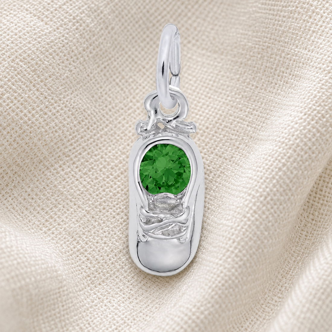 The Rembrandt Charms Baby Shoe Birthstone (May) pendant, crafted in sterling silver and featuring a green gemstone at its center, is beautifully displayed on a textured cream fabric background—an elegant keepsake charm perfect for celebrating a May birth.