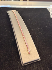 The Ryan Gems Pink Shappire Tennis Bracelet showcases a row of gemstones set in 14k rose gold, elegantly displayed on a gray cushioned stand against a black background.