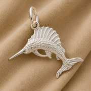 The Sailfish Charm—Sterling Silver by Rembrandt Charms, shaped like a fish with textured scales and fins, rests on a light tan fabric background.