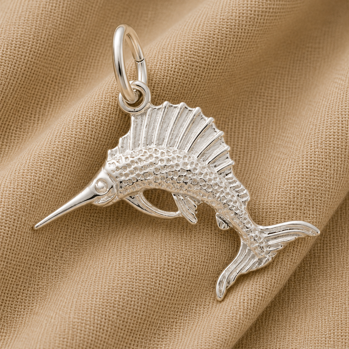 The Sailfish Charm—Sterling Silver by Rembrandt Charms, shaped like a fish with textured scales and fins, rests on a light tan fabric background.