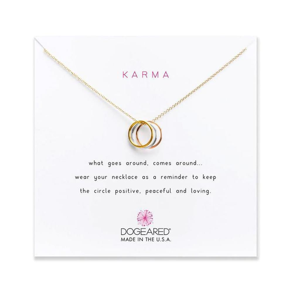 A gold necklace featuring three interlocking rings is displayed against a white background. The card behind it reads "Karma" and includes a message about keeping the circle positive, peaceful, and loving. The brand name, Dogeared, is at the bottom, labeled "Made in the U.S.A." (Karma /GP-SS).