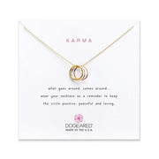 A gold necklace featuring three interlocking rings is displayed against a white background. The card behind it reads "Karma" and includes a message about keeping the circle positive, peaceful, and loving. The brand name, Dogeared, is at the bottom, labeled "Made in the U.S.A." (Karma /GP-SS).