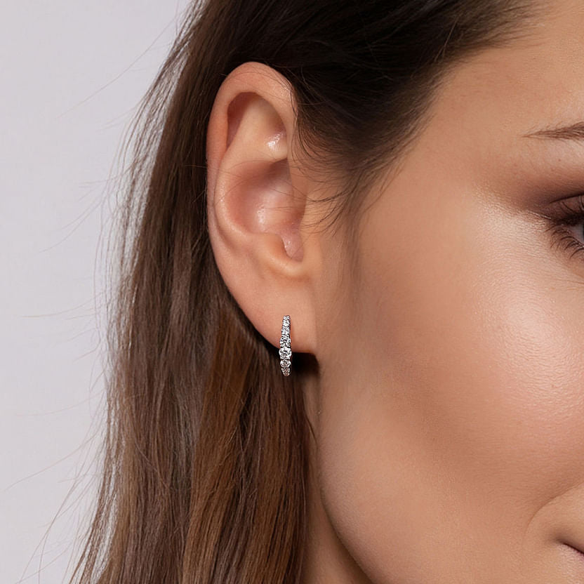 Close-up of a woman's ear wearing Gabriel & Co's 14K White Gold 13mm Classic Diamond Huggie Earrings, her brown hair tucked behind her ear and her skin smooth, highlighting the sparkling diamonds.