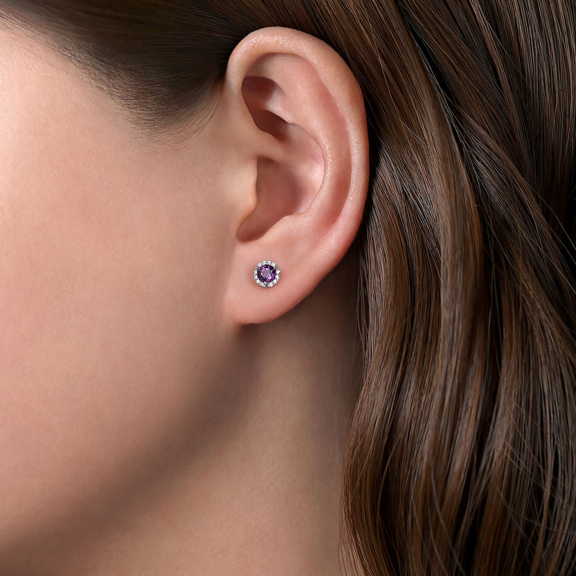 Close-up of a woman’s ear with brown hair, wearing Gabriel & Co 14K White Gold Amethyst and Diamond Halo Stud Earrings, featuring a vibrant purple amethyst center surrounded by sparkling diamonds.