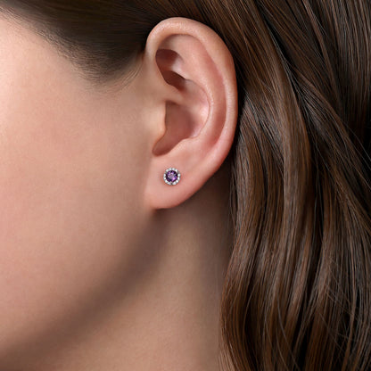 Close-up of a woman’s ear with brown hair, wearing Gabriel & Co 14K White Gold Amethyst and Diamond Halo Stud Earrings, featuring a vibrant purple amethyst center surrounded by sparkling diamonds.