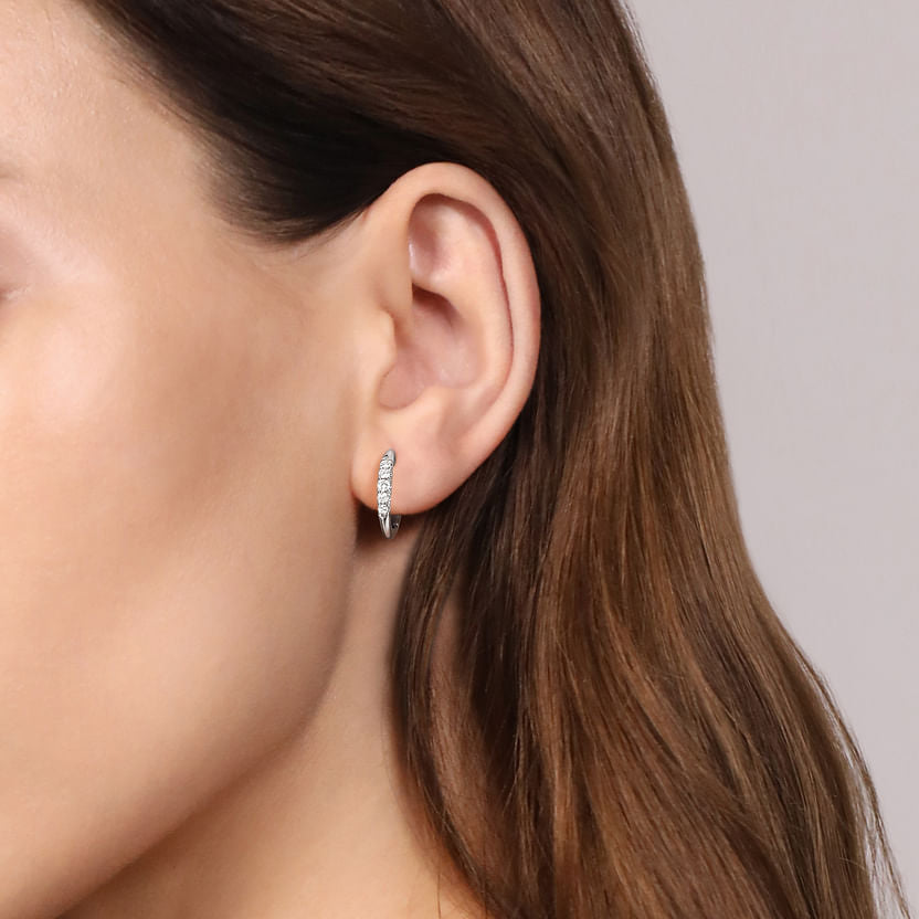 Close-up of a woman's ear with long brown hair, wearing Gabriel & Co 14K White Gold Classic Round 15mm Diamond Huggie Earrings. The background is a plain, light color.