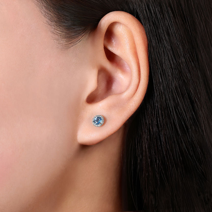Close-up of a person’s ear wearing Gabriel & Co 14K White Gold Diamond Halo Swiss Blue Topaz Stud Earrings, with the out-of-focus background highlighting the elegant gemstone and halo setting.