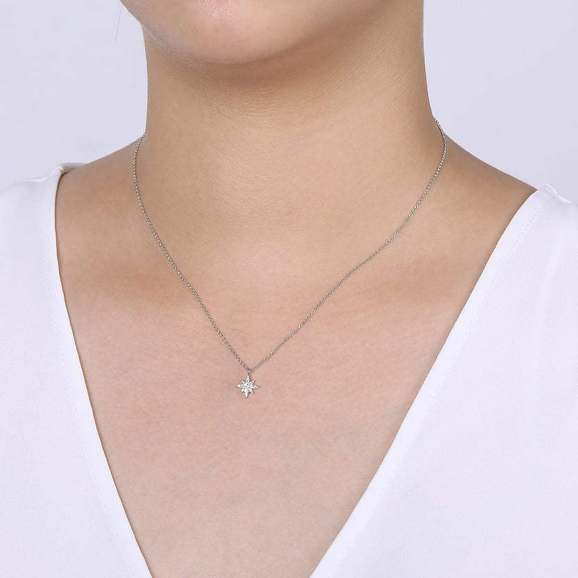 A person wearing the Gabriel & Co 14K White Gold Diamond Pave Starburst Pendant Necklace with a small starburst charm, styled with a white V-neck top against a light, plain background.