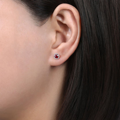 Close-up of a person with straight, dark brown hair wearing Gabriel & Co's 14K White Gold Diamond and Ruby Stud Earrings, featuring a round ruby center encircled by pavé-set natural diamonds.