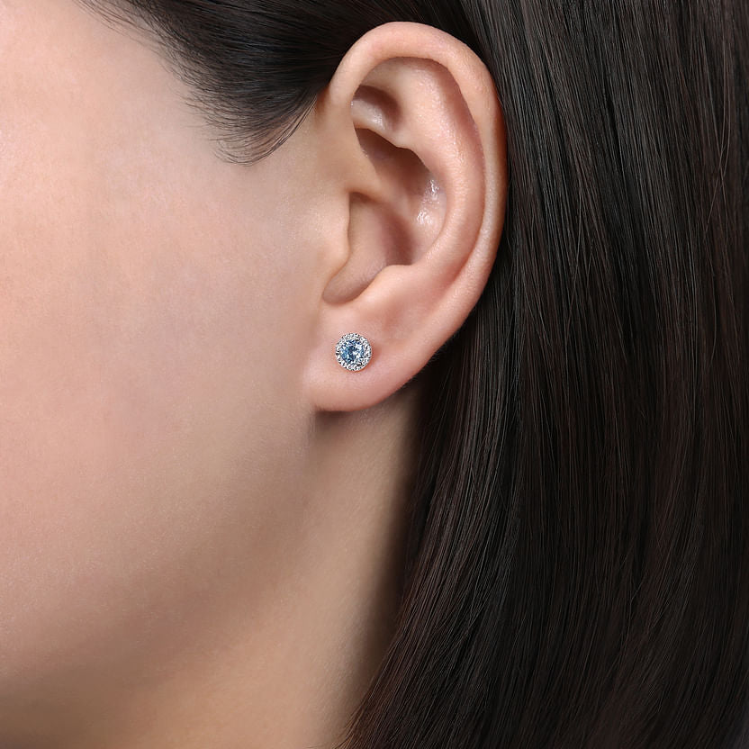 A close-up of a woman's ear showcases Gabriel & Co's 14K White Gold Halo Aquamarine and Diamond Stud Earring, featuring a blue aquamarine center surrounded by diamonds, against straight dark brown hair.
