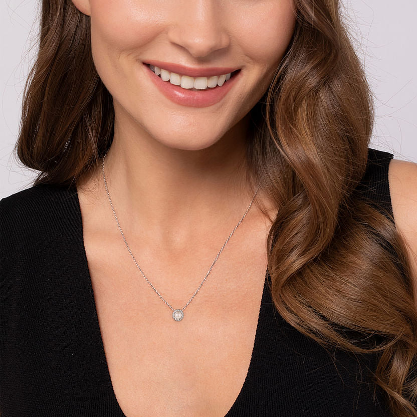 A woman with long brown hair and a light complexion smiles, wearing a black sleeveless top and the Gabriel & Co 14K White Gold Pearl and Diamond Halo Pendant Necklace, with the image focused on her neck and shoulders.