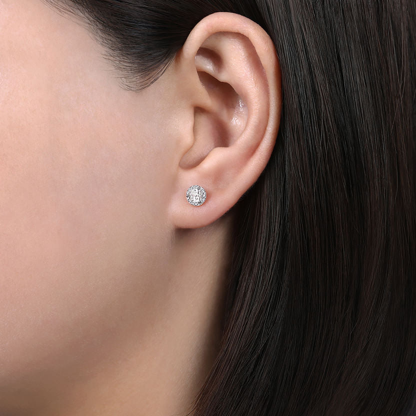 Close-up of a person’s ear wearing Gabriel & Co 14K White Gold Halo White Sapphire and Diamond Stud Earrings. Their straight, dark brown hair is tucked back, highlighting the sparkling accessory against a neutral background.