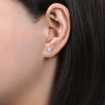 Close-up of a person’s ear wearing Gabriel & Co 14K White Gold Halo White Sapphire and Diamond Stud Earrings. Their straight, dark brown hair is tucked back, highlighting the sparkling accessory against a neutral background.