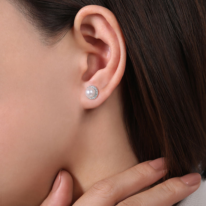 Close-up of a woman's ear adorned with Gabriel & Co 14K White Gold Round Diamond Halo Pearl Stud Earrings. Her neatly manicured hand touches her neck, and brown hair is tucked behind her ear.