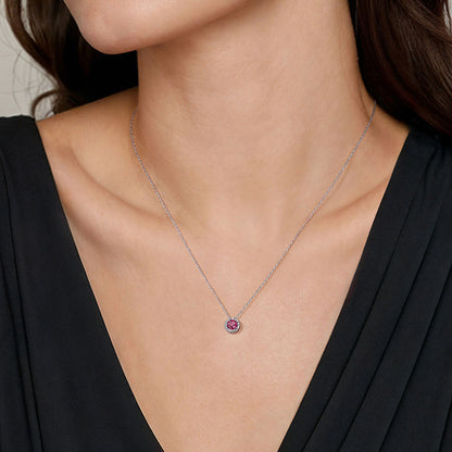 A woman in a black V-neck dress wears the Gabriel & Co 14K White Gold Ruby and Diamond Halo Pendant Necklace, showcasing elegant July birthstone jewelry at the center of her chest.