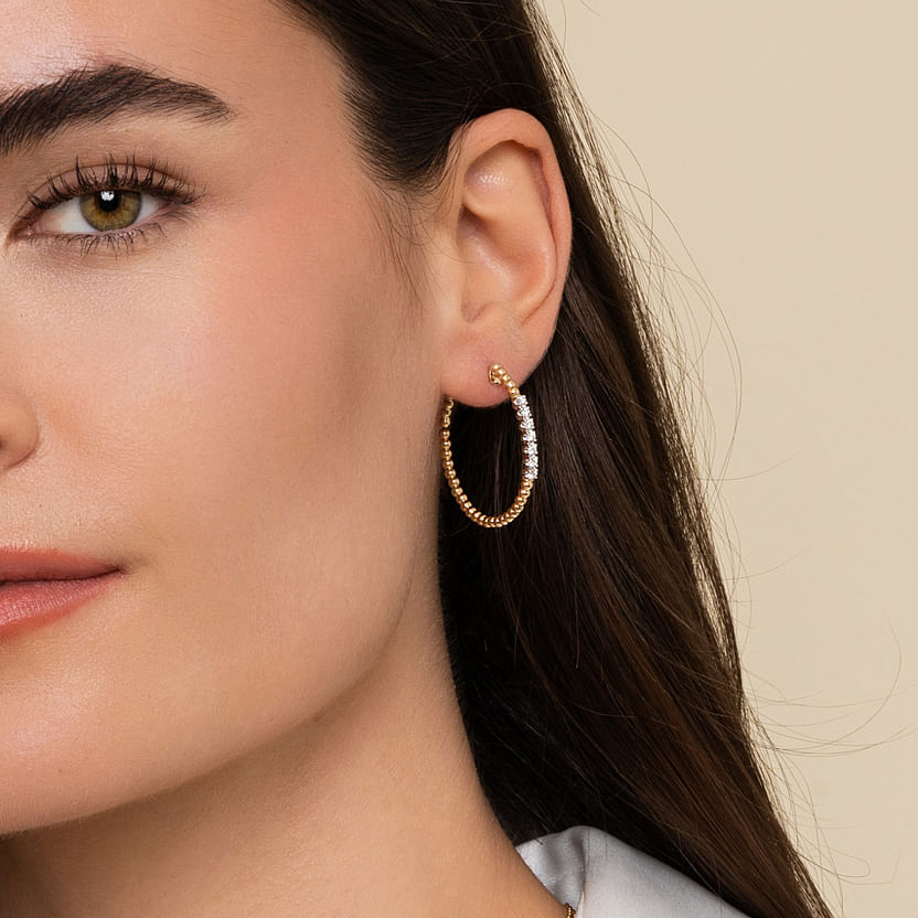 Close-up of a woman with straight brown hair wearing Gabriel & Co 14K Yellow Gold 30mm Bujukan Diamond Classic Hoop Earrings. She has on a light top, and the background is plain beige. Her left ear is clearly visible, showcasing the earrings.