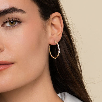 Close-up of a woman with straight brown hair wearing Gabriel & Co 14K Yellow Gold 30mm Bujukan Diamond Classic Hoop Earrings. She has on a light top, and the background is plain beige. Her left ear is clearly visible, showcasing the earrings.
