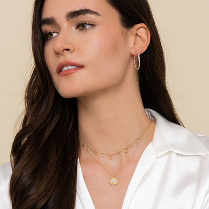 A woman with long brown hair in a white satin blouse wears Gabriel & Co layered necklaces and 14K yellow gold 30mm Bujukan Diamond Classic Hoop Earrings, looking slightly to the side against a plain background.