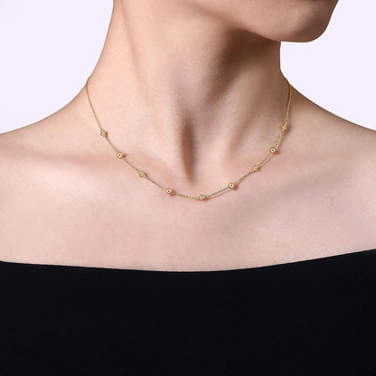 A person in an off-the-shoulder black top wears the 14K Yellow Gold Bujukan Bead Station Necklace by Gabriel & Co. against a plain, light background, highlighting this elegant piece from Avonlea Jewelers.