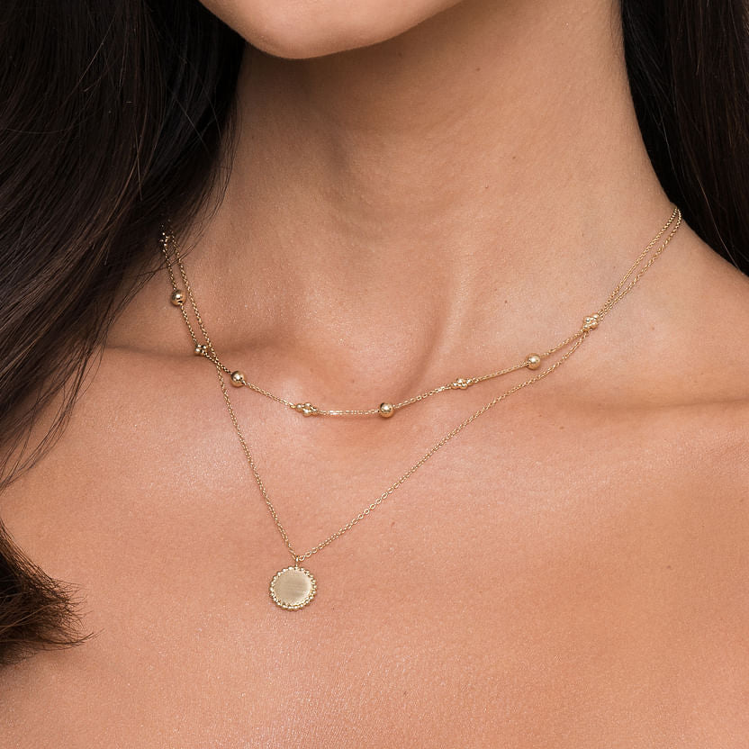 A woman wears two delicate gold necklaces: the 14K Yellow Gold Bujukan Bead Station Necklace by Gabriel & Co and another with a round, flat pendant. Both rest gracefully on her bare neck and collarbone.