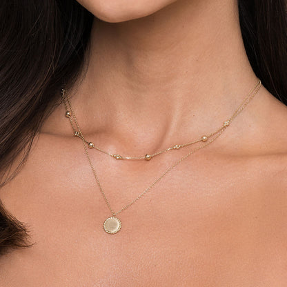 A woman wears two delicate gold necklaces: the 14K Yellow Gold Bujukan Bead Station Necklace by Gabriel & Co and another with a round, flat pendant. Both rest gracefully on her bare neck and collarbone.