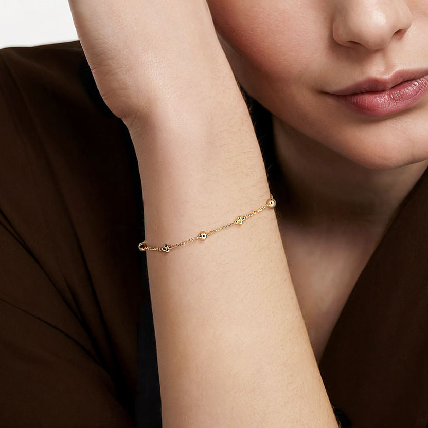 A woman in a brown top showcases the 14K Yellow Gold Bujukan Beads Station Bracelet by Gabriel & Co, with her arm resting on a surface. Avonlea Jewelers offers this timeless piece for elegant style.