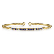The Gabriel & Co 14K Yellow Gold Bujukan Diamond and Blue Sapphire Split Bangle features beaded detailing, a central bar with alternating sapphires and diamonds, and two round gold end caps.