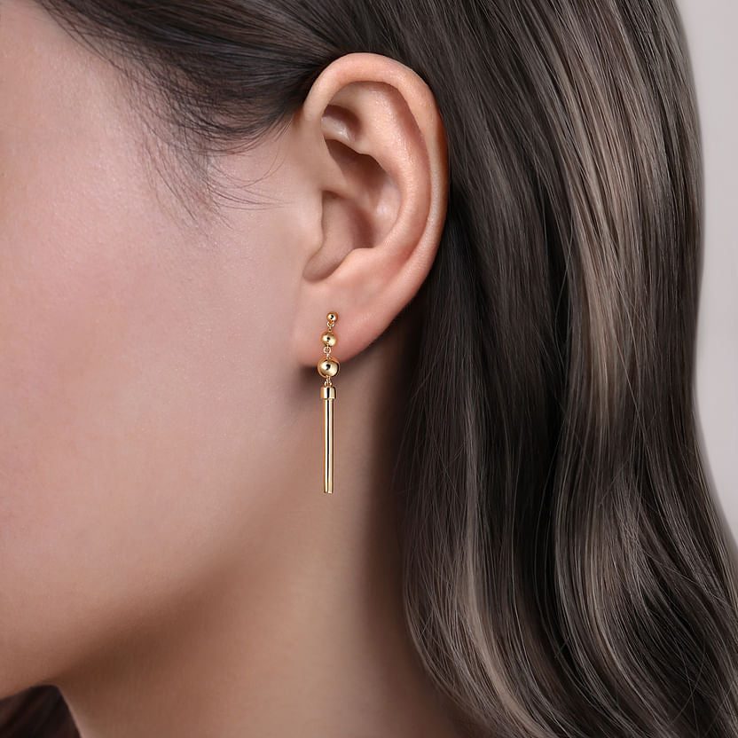 A close-up of a person with straight brown hair wearing three 14K yellow gold studs and a long, thin Gabriel & Co 14K Yellow Gold Bujukan Drop Earring.