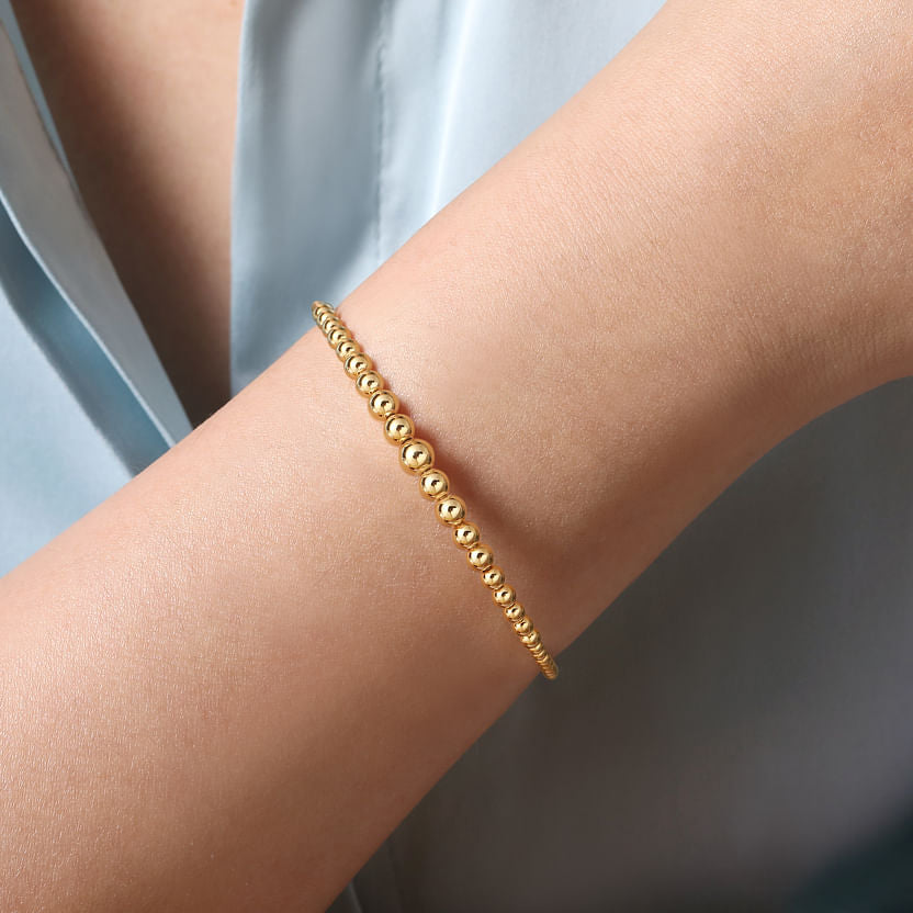 A close-up of a person wearing a 14K Yellow Gold Bujukan Open Bangle by Gabriel & Co on their wrist, with the sleeve of a light blue shirt visible in the background.