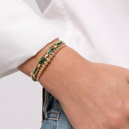 A person in a white long-sleeve shirt and blue jeans wears the Gabriel & Co 14K Yellow Gold Bujukan Open Bangle, featuring green stones and gold beads for an elegant yet modern minimalist touch.