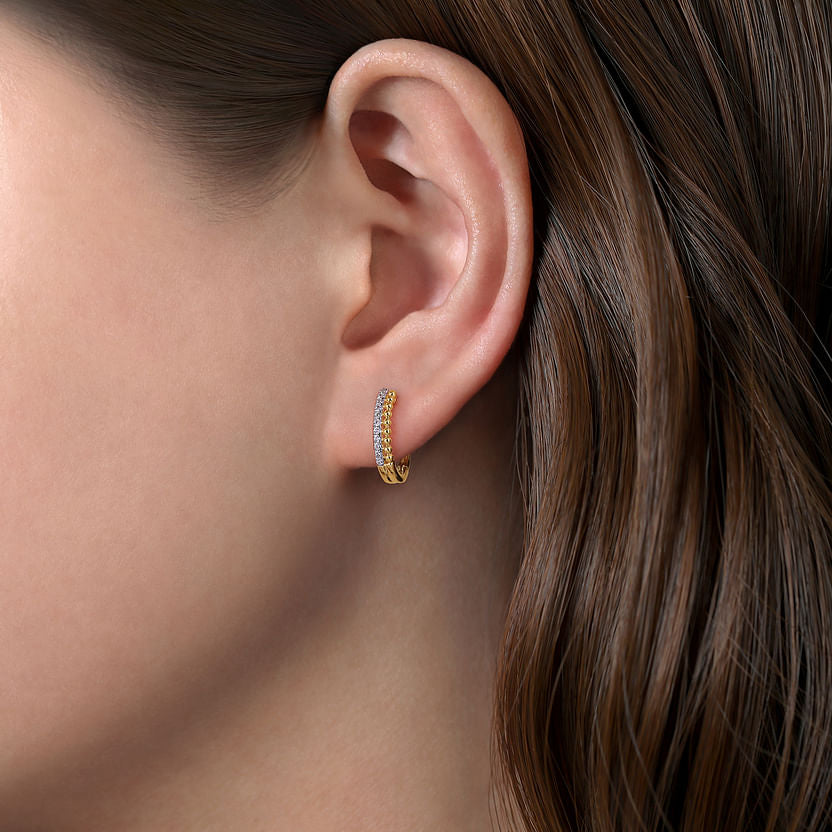 Close-up of a woman's ear adorned with Gabriel & Co's 14K Yellow Gold Bujukan Pave 10mm Diamond Huggie Earrings, shimmering with clear stones. The sparkling earring stands out against her smooth brown hair and a softly blurred background.