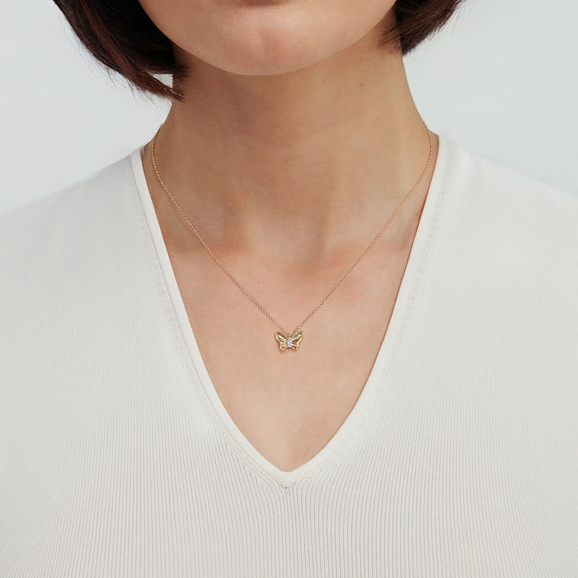 A woman in a white V-neck top is shown from the shoulders down, wearing the Gabriel & Co 14K Yellow Gold Butterfly Necklace with a diamond cut finish and graduating diamonds.