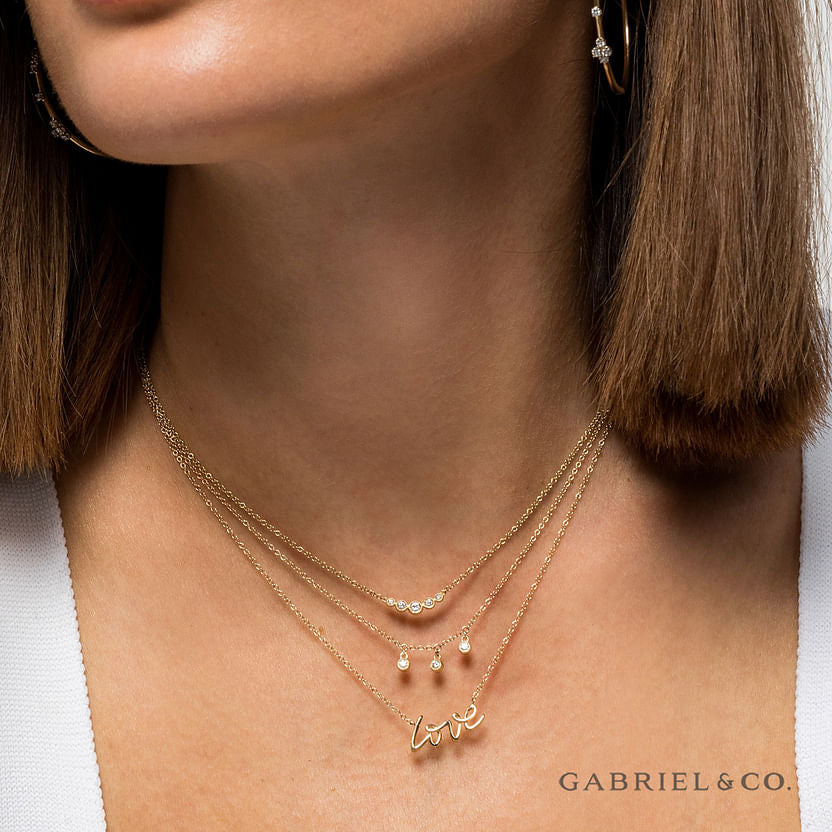 A woman wears three layered gold necklaces: the 14K Yellow Gold Curved Diamond Bar Necklace by Gabriel & Co, one with vintage charms, and a "love" pendant. The Gabriel & Co logo appears in the corner.