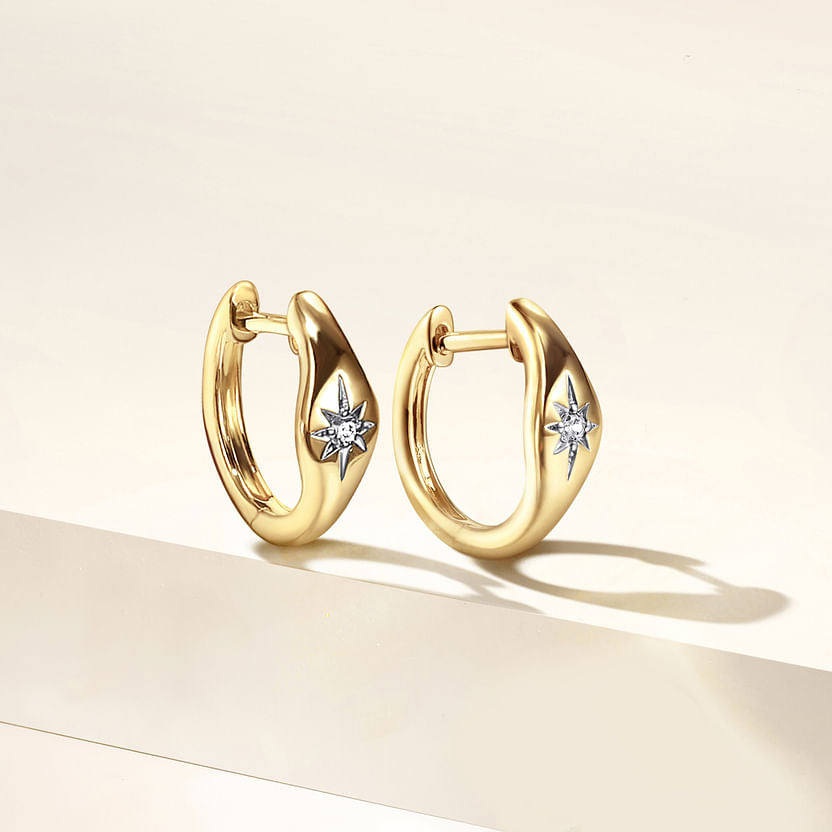A pair of 14K Yellow Gold Diamond Huggie Earrings by Gabriel & Co, featuring star-shaped diamond accents, displayed on a cream-colored surface with soft lighting.