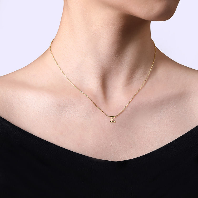 A person wearing the Gabriel & Co 14K Yellow Gold E Initial Necklace, featuring a small "E" pendant, styled with a black off-the-shoulder top against a light background.