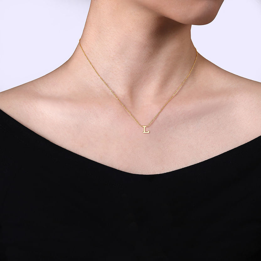 A person wears the Gabriel & Co 14K Yellow Gold L Initial Necklace, featuring a delicate "L" pendant. Shown from the shoulders up against a light background, their black off-shoulder top highlights this elegant piece.