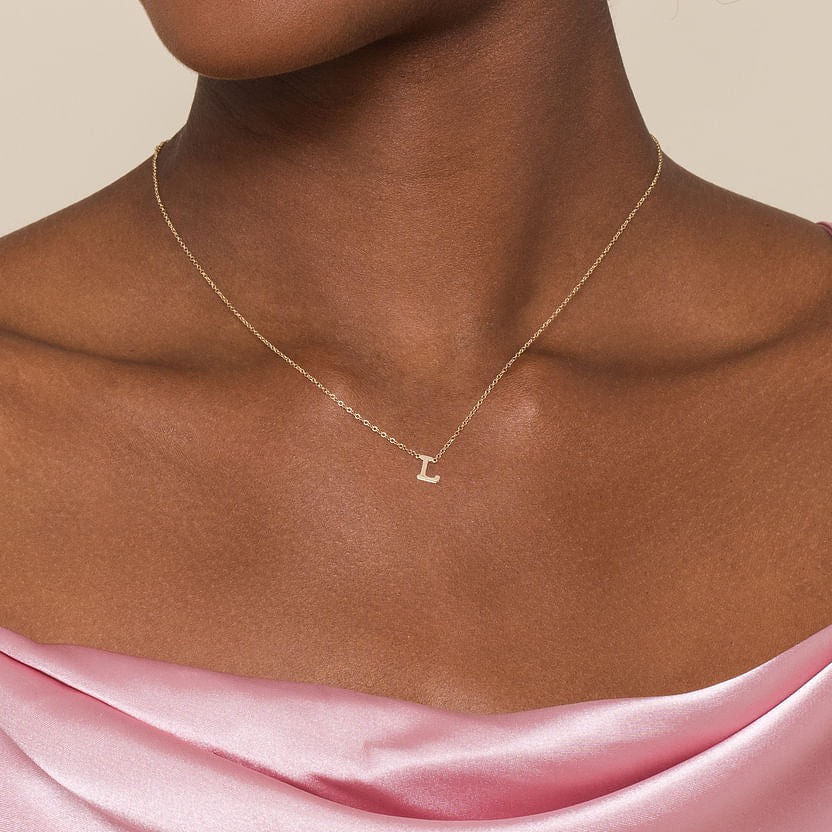 A woman wears a pink off-the-shoulder satin top and the Gabriel & Co 14K Yellow Gold L Initial Necklace, featuring a small letter "L" pendant, standing against a plain, neutral background.