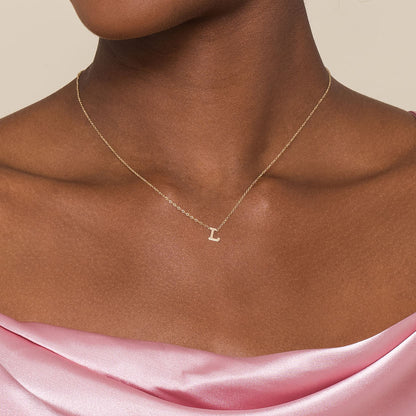 A woman wears a pink off-the-shoulder satin top and the Gabriel & Co 14K Yellow Gold L Initial Necklace, featuring a small letter "L" pendant, standing against a plain, neutral background.