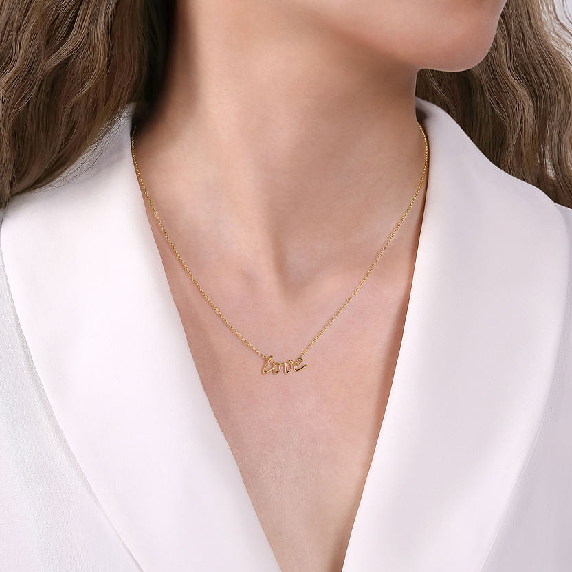 A woman, her wavy hair visible and only her lower face and neck shown, wears a white blouse and the 14K Yellow Gold Love Necklace by Gabriel & Co.
