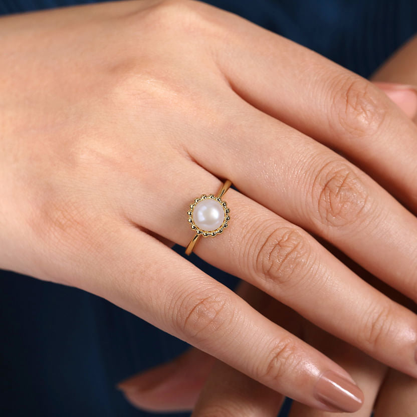 A hand showcases the Gabriel & Co 14K Yellow Gold Pearl Ring with Bujukan beaded halo, featuring a cultured pearl and accent stones on the ring finger against a dark blue background.