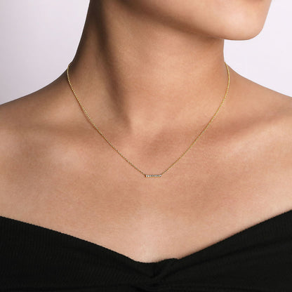 A person wears the Gabriel & Co 14K Yellow Gold Petite Pave Diamond Bar Necklace with a black off-the-shoulder top against a plain, light background. The necklace features a small, horizontal diamond bar pendant.
