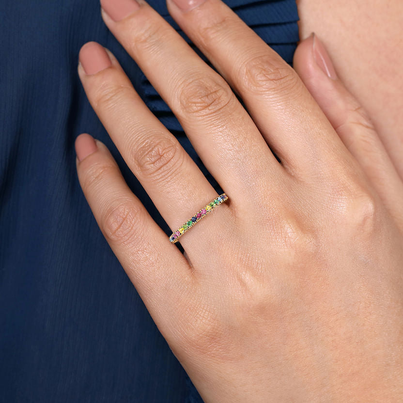 A hand with manicured nails wears a Gabriel & Co 14K Yellow Gold Rainbow Color Stone Stackable Ring, its multicolor gems gleaming atop the band as the hand rests gently against dark blue, pleated fabric.