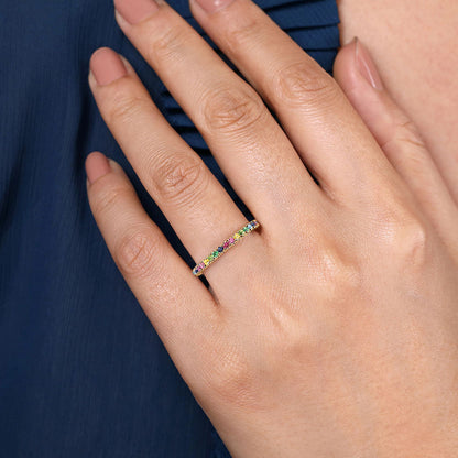 A hand with manicured nails wears a Gabriel & Co 14K Yellow Gold Rainbow Color Stone Stackable Ring, its multicolor gems gleaming atop the band as the hand rests gently against dark blue, pleated fabric.