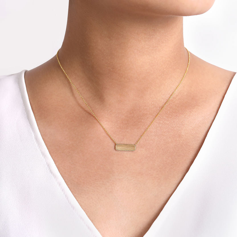 A person shown from the shoulders up wears the Gabriel & Co 14K Yellow Gold Rectangular ID Pendant Necklace with Twisted Rope Frame over a white V-neck top, embodying personalized elegance against a neutral light background.